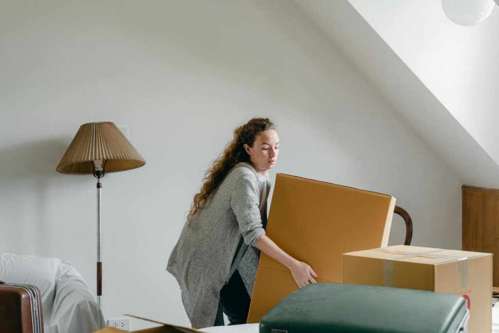Slim female in cardigan taking big rectangular cardboard box while standing near suitcases and sofa in attic style living room in daylight