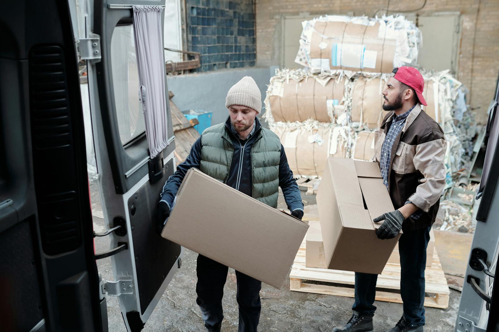 Two male workers loading cardboard boxes in a warehouse with bundled paper in the background.