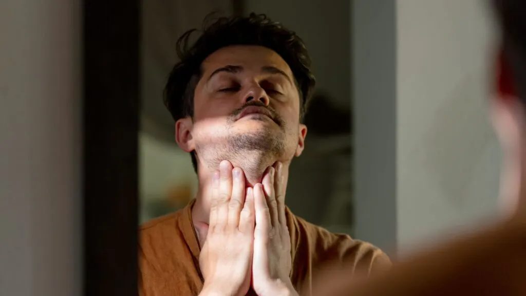 A man standing in front of a mirror gently massaging the front of his neck with both hands.