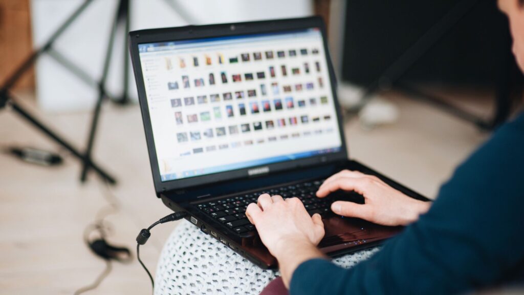 A person sitting down, viewed from over the shoulder, typing on a black laptop whose screen displays a large gallery of small image thumbnails.
