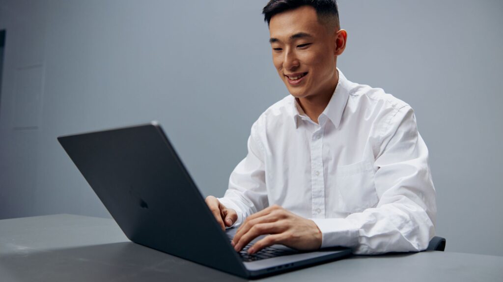 A smiling young Asian man in a white button-down shirt sits at a desk, happily typing on a modern dark gray laptop with a neutral gray background.