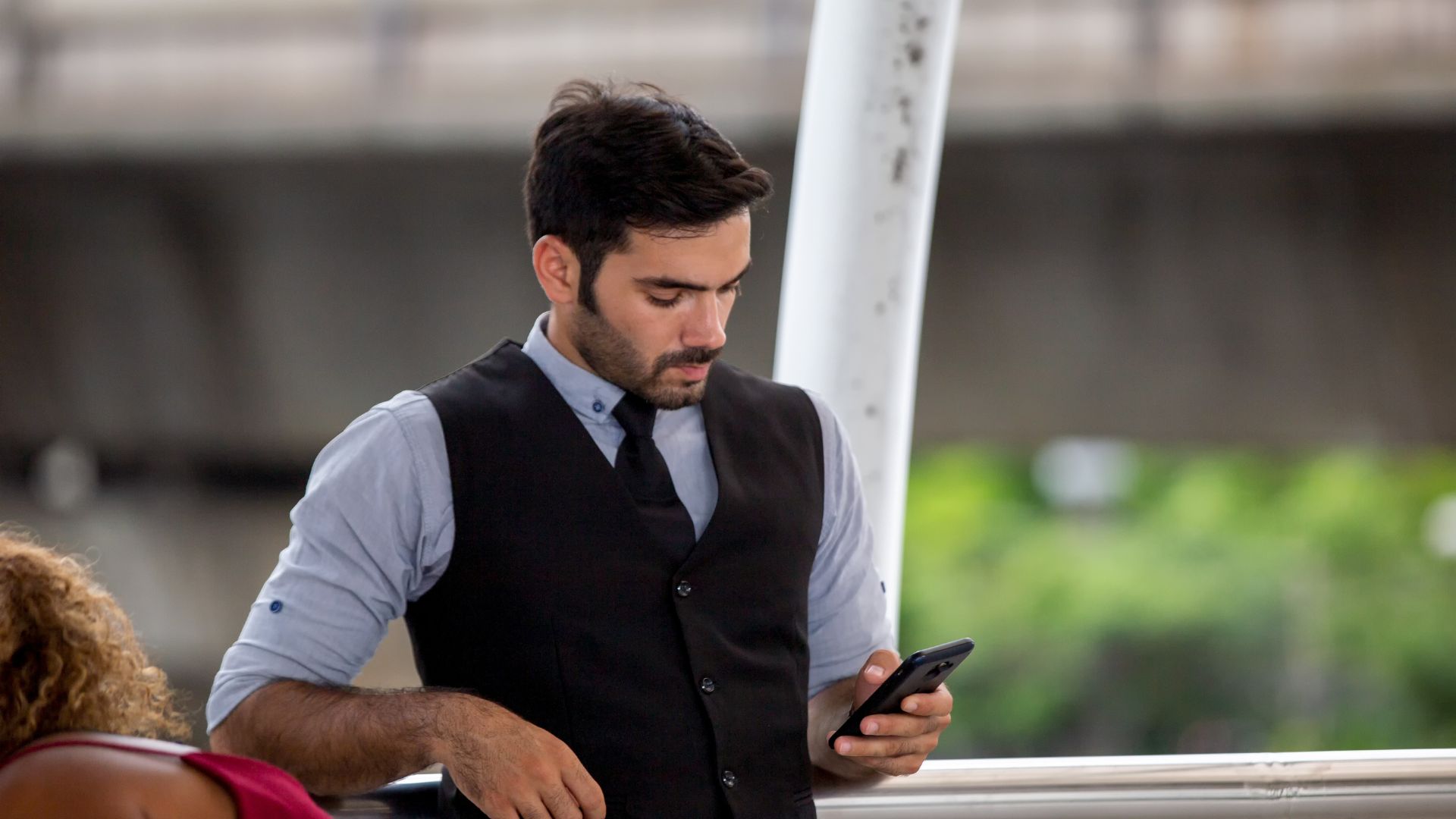 A formally dressed man with a beard, wearing a vest and tie, stands outdoors leaning on a railing while focused on reading content on his smartphone.