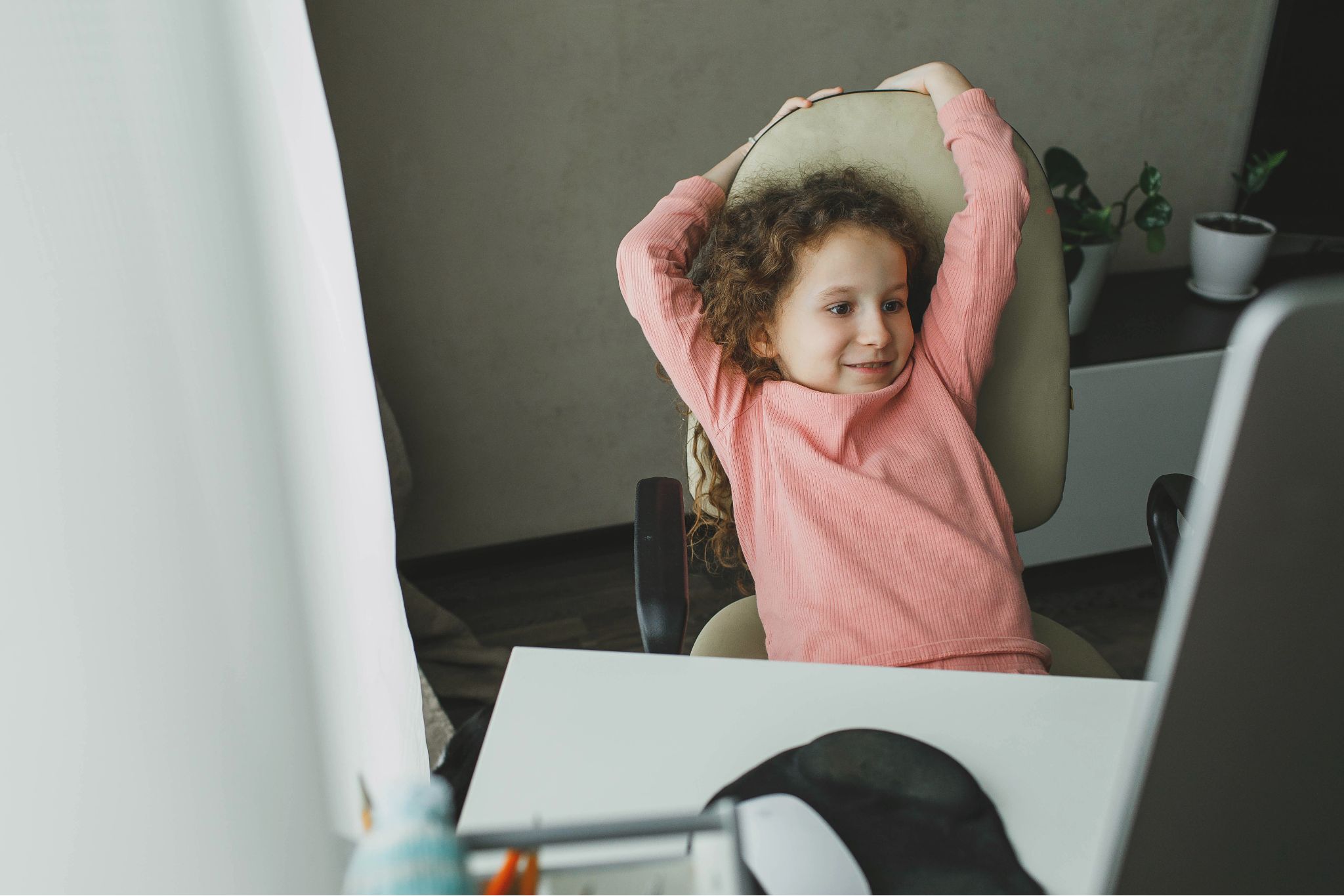 Happy child with curly hair stretching in a chair in front of a computer monitor.