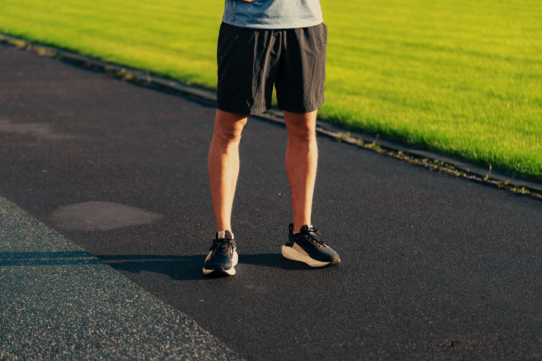 A person is standing on an outdoor running track, wearing athletic shorts and running shoes. The focus is on their legs, with bright green grass visible in the background.