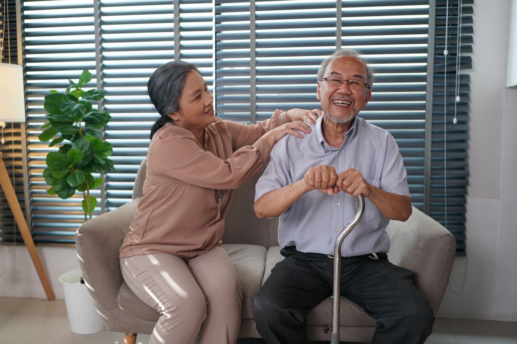 Design sem nome – 2025-10-03T213338.994 Happy senior Asian couple sitting on a couch, the woman is gently massaging her husband's shoulder.