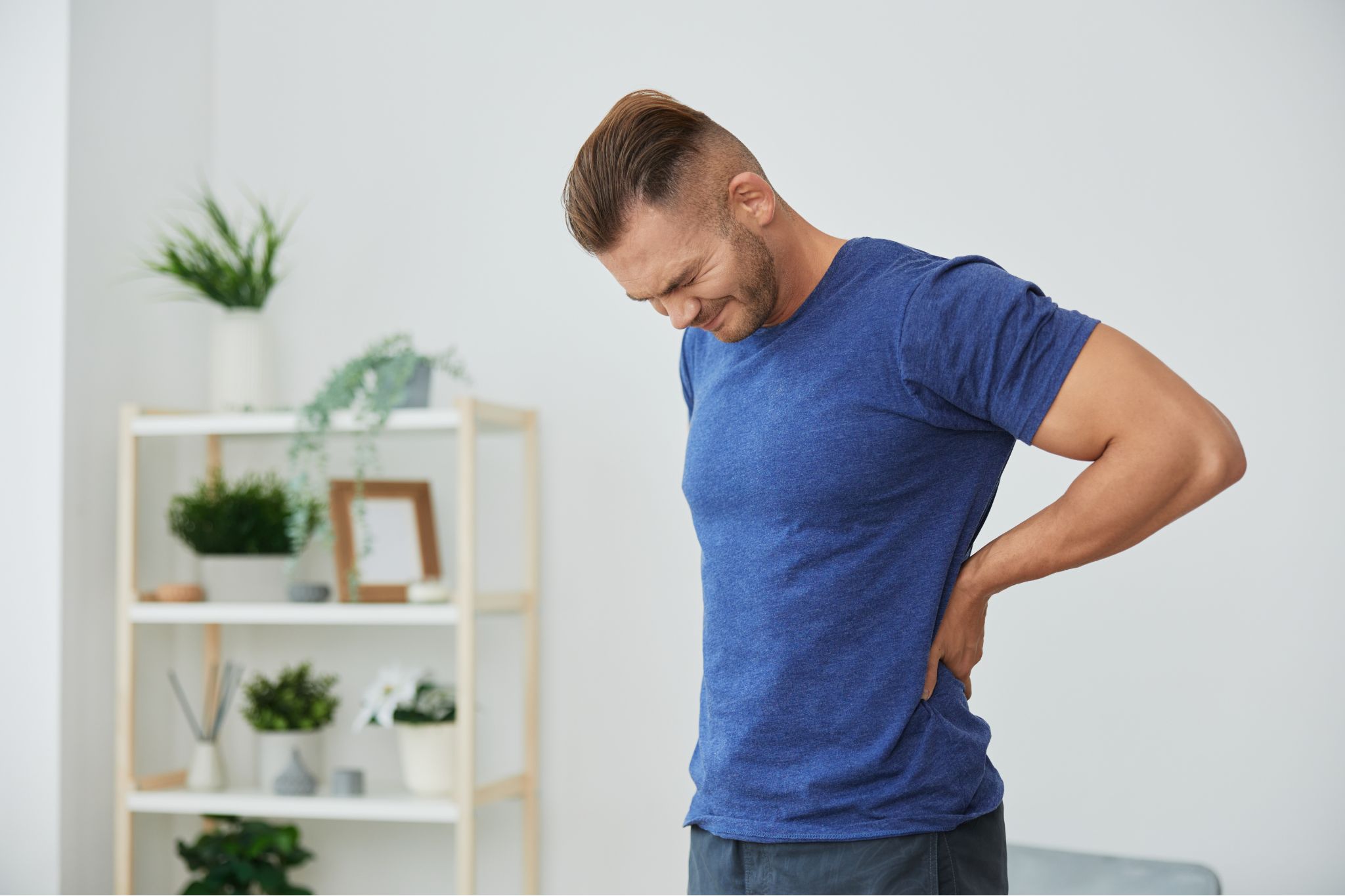 Young man in a blue t-shirt standing indoors, holding his lower back in pain.