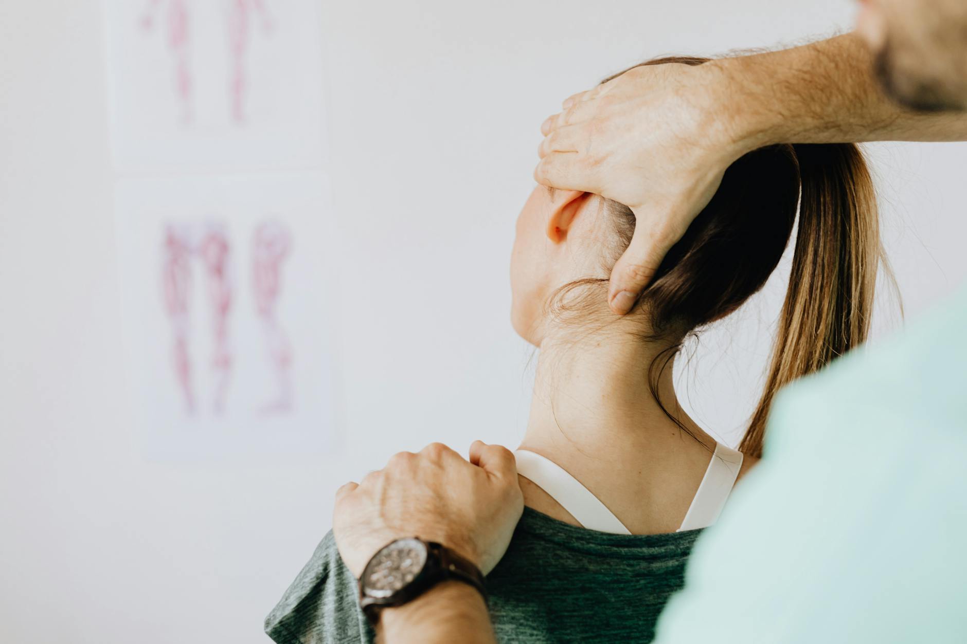 Back view of crop faceless bearded orthopedist in wristwatch checking up neck of anonymous lady while standing in front of wall with paper drawings representing body anatomy